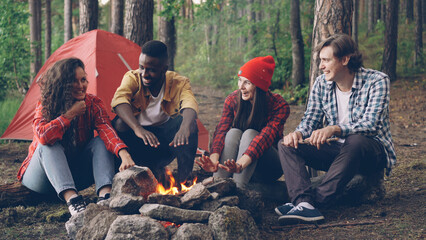 Young men and women happy travelers are sitting around fire, telling stories and laughing, handsome...