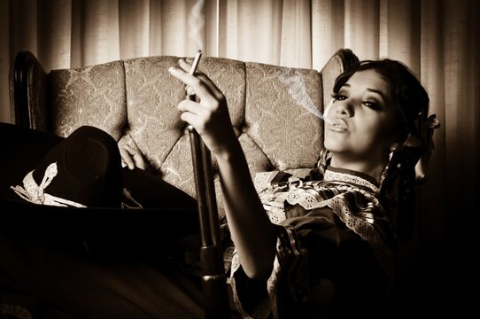 Retrato de una mujer modelo adolescente sentada mientras viste un traje típico de Adelita de la Revolución Mexicana, un sombrero de charro, un rifle, y fumando un cigarro