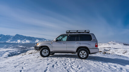 Fototapeta na wymiar The SUV is standing on a snow-covered high-altitude plateau. Footprints are visible all around. A mountain range against a blue sky. Copy space. Altai