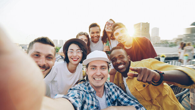 Handsome young man in cap is looking at camera to take selfie, people are having fun, making funny faces and gestures and laughing at rooftop party.