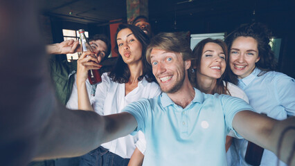 Group of happy colleagues are taking selfie together, young man is holding camera and posing, his coworkers are holding drinks in bottles, laughing and looking at camera. - obrazy, fototapety, plakaty