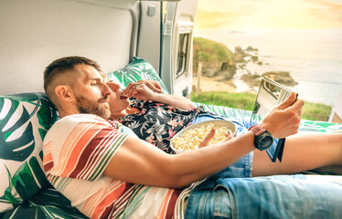 Young couple watching a movie on the tablet eating popcorn lying on bed of their camper van with...