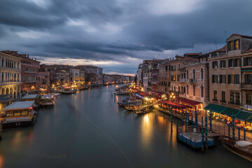 Fototapeta na wymiar Evening over the Grand Canal from Rialto Bridge in Venice, Italy. The sky is cloudy and the canal is lit up