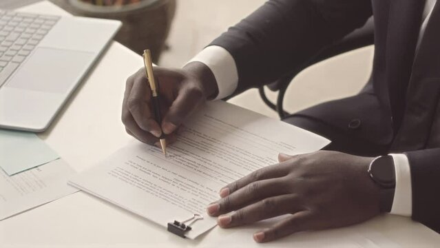 Cropped of unrecognizable African American businessman in stylish black suit putting signature in contract sitting at office desk