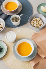 Top view of bowl with pumpkin soup. Near pumpkin seeds in a bowl, sour cream, crackers and second soup bowl. The table is covered with a linen tablecloth, with yellow and blue placemats.