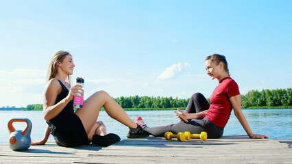 Two beautiful, athletic blonde girls, women have rest after training outdoors, drink water, talk, laugh. Lake, river, blue sky and forest in the background, summer sunny day. High quality photo