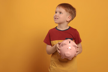 Fototapeta na wymiar Cute little boy with ceramic piggy bank on orange background, space for text
