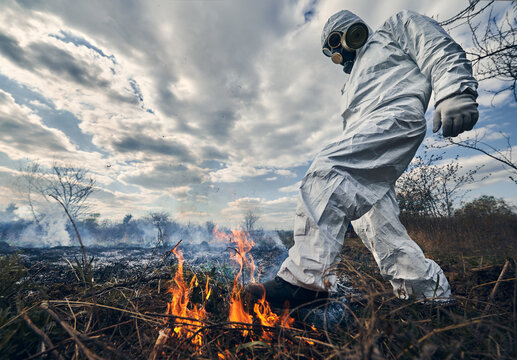 Firefighter ecologist fighting fire in field. Man in protective radiation suit and gas mask near burning grass with smoke. Natural disaster concept.