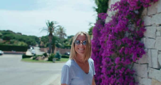 Model on travel video, smile. Happy woman spins around and walks along a wall overgrown with blossom flowers. Attractive and cute smile. Genuine emotions. Freedom and happiness summer vacation vibe. 