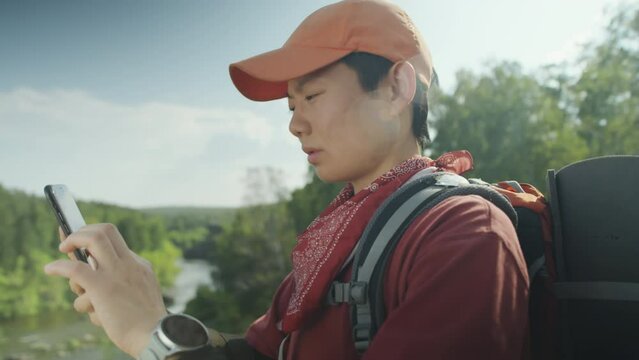 Medium close up shot of Asian female tourist with backpack standing in national park and using mobile phone and smartwatch for navigation during hike