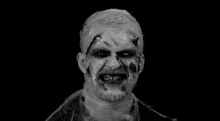 Fototapeta premium Portrait of sinister man in carnival costume of Halloween crazy zombie with bloody wounded scars face trying to scare isolated on black room. Horror theme of cosplay wounded undead, beast, monster