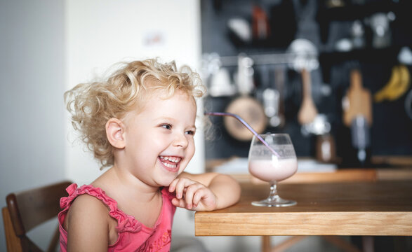 Emotional portrait of a child girl with curly hair at home in the kitchen with a glass of smoothie. Healthy breakfast and fun.
