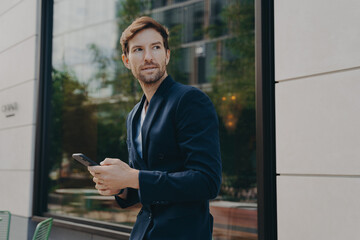 Businessman in dark suit holding smartphone and looking aside while walking city street