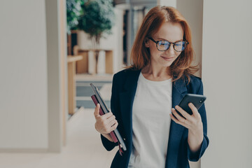 Elegant smiling woman office worker in eyeglasses using modern mobile phone in coworking space