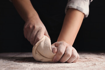 Man kneading dough at wooden table on dark background, closeup