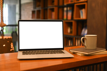 Mock up laptop computer, books and coffee cup on wooden table against blurred bookshelf background