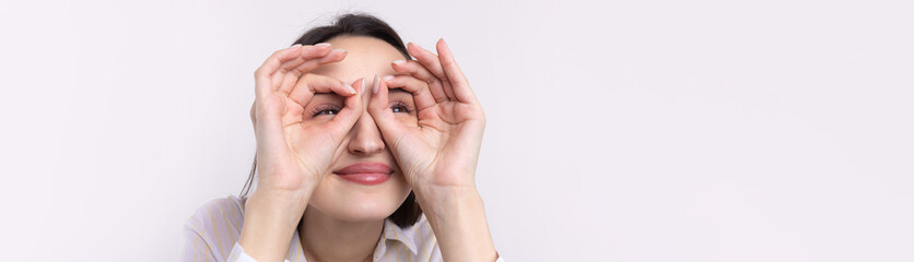 Close up portrait of attractive quirky young woman making binoculars with hands showing ok gesture on white studio background.