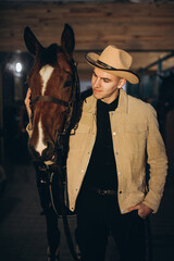 A handsome young cowboy is holding a horse by the leash. A man poses with a horse in a stable.