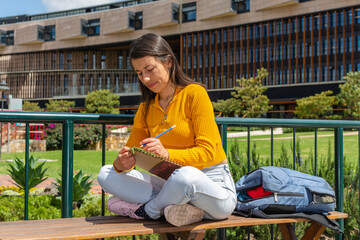 Young Hispanic female student sitting on a college campus bench wearing a yellow sweater while...