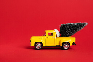 Yellow retro toy pickup carrying a Christmas tree on red background. Christmas and New Year...