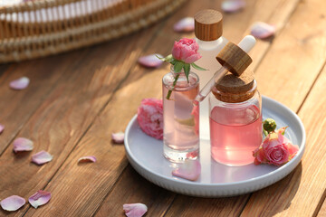 Bottles of rose essential oil and flowers on wooden table, space for text