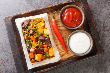 Turkish kebab with potatoes, green peas, onions and carrots wrapped in paper and baked close-up on a wooden tray on the table. Horizontal top view from above