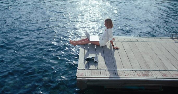 Young business woman sit on edge of pier and work on laptop. Lay down and relax, tired of working. Out of office, work on holiday. Content creator work in resort and vacation