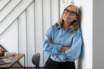 portrait of a successful Middle aged older business woman leaning against the wall in the office,...