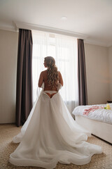 view from the back. a blonde bride wears a white dress by the bedroom window.