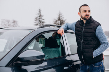 Handsome man in warm jacket standing by car covered with snow