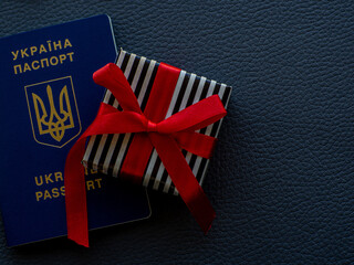Small gift in a stripped black and white wrapper and with red ribbons and passport of Ukraine on the dark background. Travel for Ukrainians, a trip as a gift concept. Copy space for your text.