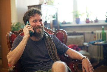 Reggae hippie man sitting in his living room, having a chat on his cell phone, sitting in a nice...