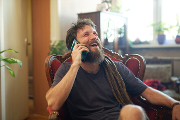 Reggae hippie man sitting in his living room, having a chat on his cell phone, sitting in a nice...