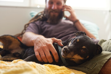 A bearded rastafarian hippie man petting his dog while talking on the phone, laying in his bed
