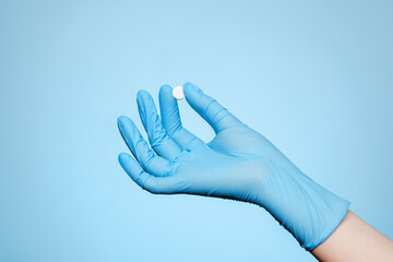 Woman's hand in a blue medical glove holding white round pill on a blue background. Close up.