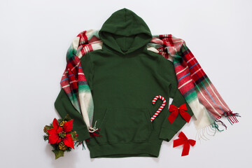 Close up green blank template hoodie copy space. Christmas Holiday concept. Top view mockup hoodie, scarf, hat. Red holidays decorations white background. Happy New Year accessories. Selective focus