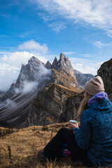Fototapeta premium A relaxed girl with a cup of tea enjoying the view sits in front of the Seceda mountain which is partially hidden in clouds. Autumn colours. Focus on a mountain.