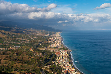 Top view of the Sicilian coast. In the background, buildings along the coast, mountains, the Mediterranean Sea and clouds in the sky