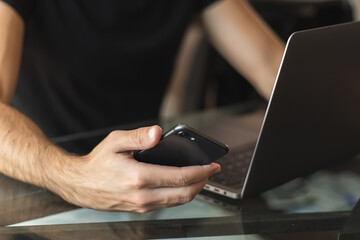 Male on black t-shirt working on his laptop while holding phone, comminication. Remote work place. Close up view.