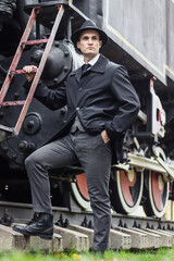 Fototapeta na wymiar Men's photo session in a classic plaid suit and hat against the backdrop of an old steam locomotive.