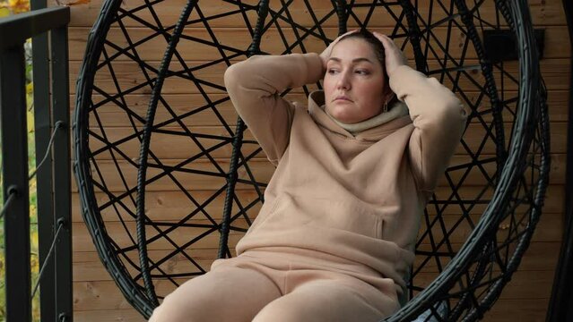 Sorrowful brunette woman wearing sports suit sits in black hanging cocoon chair on terrace. Person wipes face with palms from grief and put on hood