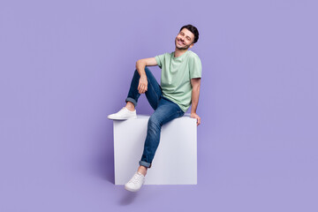Full length photo of nice young man sitting white copyspace cube cheerful dressed stylish gray clothes isolated on purple color background