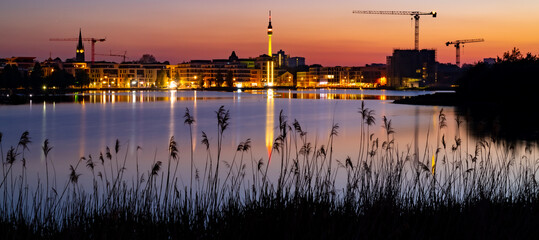 Dortmund wide angle nighttime panorama. German city with downtown skyline, modern buildings  and pond. Evening sky with clouds and reflection in Phoenix lake at blue hour with colorful illumination - obrazy, fototapety, plakaty