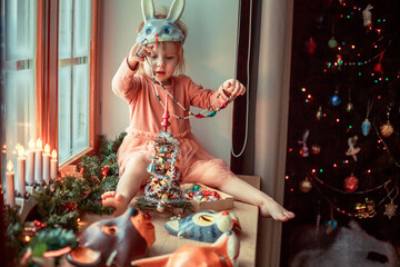 Cute little girl is dressing up a tiny Christmas tree and trying on New Year's animal masks. Image with selective focus and toning. 