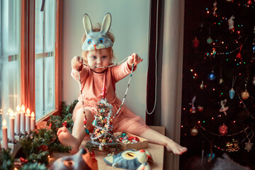 Cute little girl is dressing up a tiny Christmas tree and trying on New Year's animal masks. Image with selective focus and toning. 