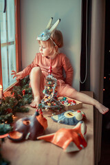 Cute little girl is dressing up a tiny Christmas tree and trying on New Year's animal masks. Image with selective focus and toning. 
