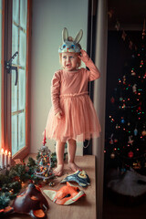 Cute little girl is dressing up a tiny Christmas tree and trying on New Year's animal masks. Image with selective focus and toning. 