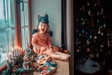 Cute little girl is dressing up a tiny Christmas tree and trying on New Year's animal masks. Image with selective focus and toning. 