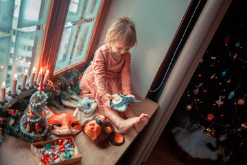 Cute little girl is dressing up a tiny Christmas tree and trying on New Year's animal masks. Image with selective focus and toning. 