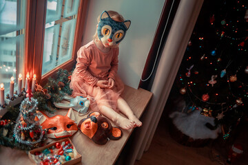 Cute little girl is dressing up a tiny Christmas tree and trying on New Year's animal masks. Image with selective focus and toning. 